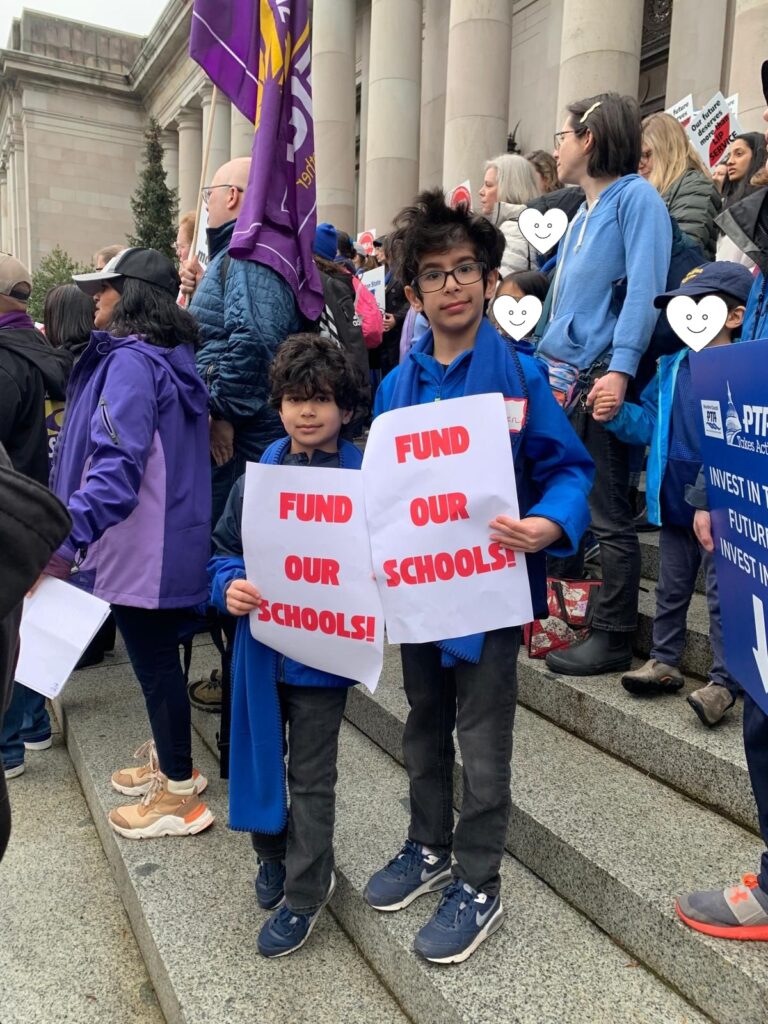 Two boys in blue scarves hold signs with "FUND OUR SCHOOLS" printed in bold red lettering, while standing as part of a crowd of people at the Washington State Capitol campus in Olympia, on Feb. 17, 2025, as part of Washington State PTA's Focus on Advocacy Day.