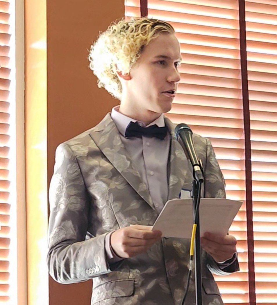 A teenager with curly blond hair and a gray floral print blazer and a black bowtie stands before a microphone holding a printed speech.